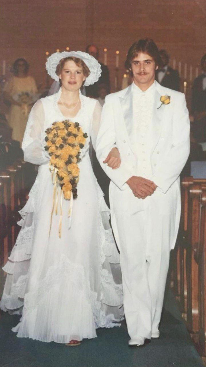 1978 Wedding Dresses and 4 Timeless Styles to Know 1 A bride in a tiered lace wedding dress and hat walks with her groom down an aisle.