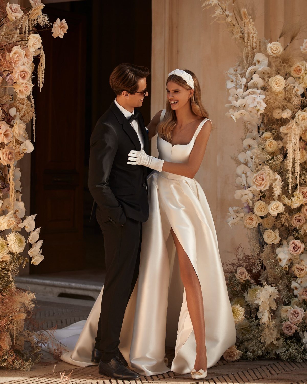 A bride in a white satin wedding dress and a groom in a black tuxedo standing amidst floral arrangements.