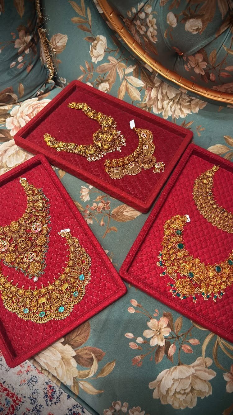 Ornate gold necklaces with green gemstones displayed on red velvet trays atop floral fabric.