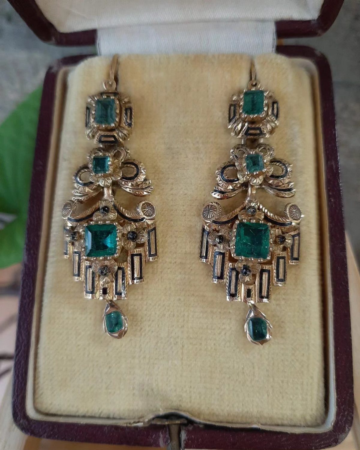 Ornate gold drop earrings with green emerald gemstones resting inside a velvet jewelry box.