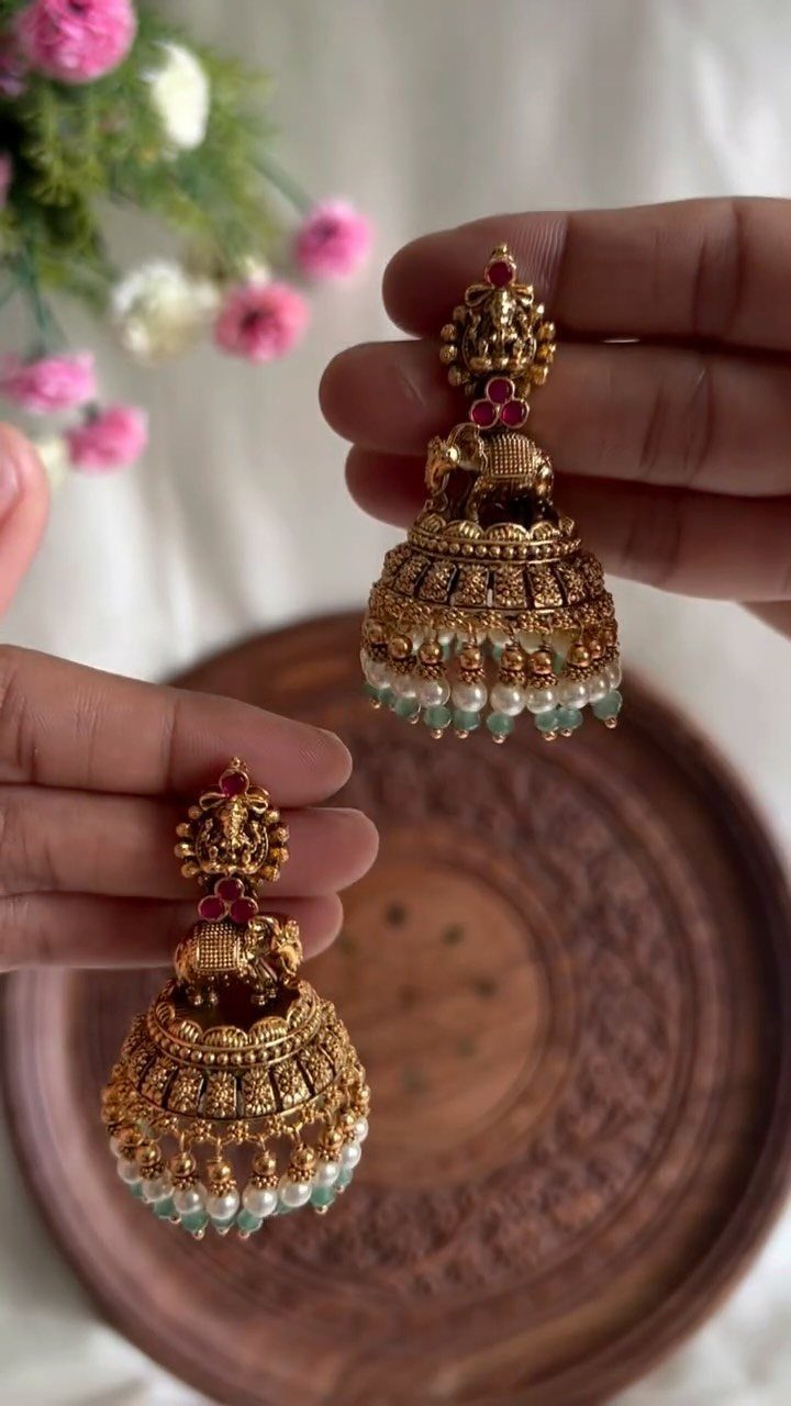 Hands holding a pair of gold jhumka earrings with red stones and hanging pearls.