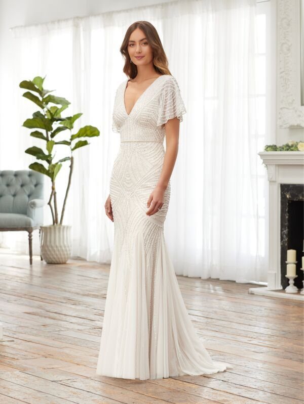 Art Deco Wedding Dress: 11 Styles From Beaded Trains to Choker Accents 3 A woman wearing a beaded white mermaid wedding dress with flutter sleeves in a bright room.