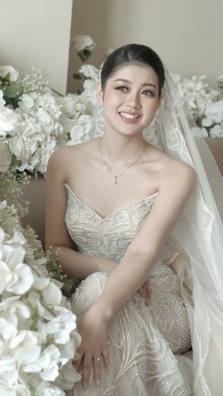 Art Deco Wedding Dress: 11 Styles From Beaded Trains to Choker Accents 10 A smiling woman in a beaded strapless wedding dress with a white veil and floral background.