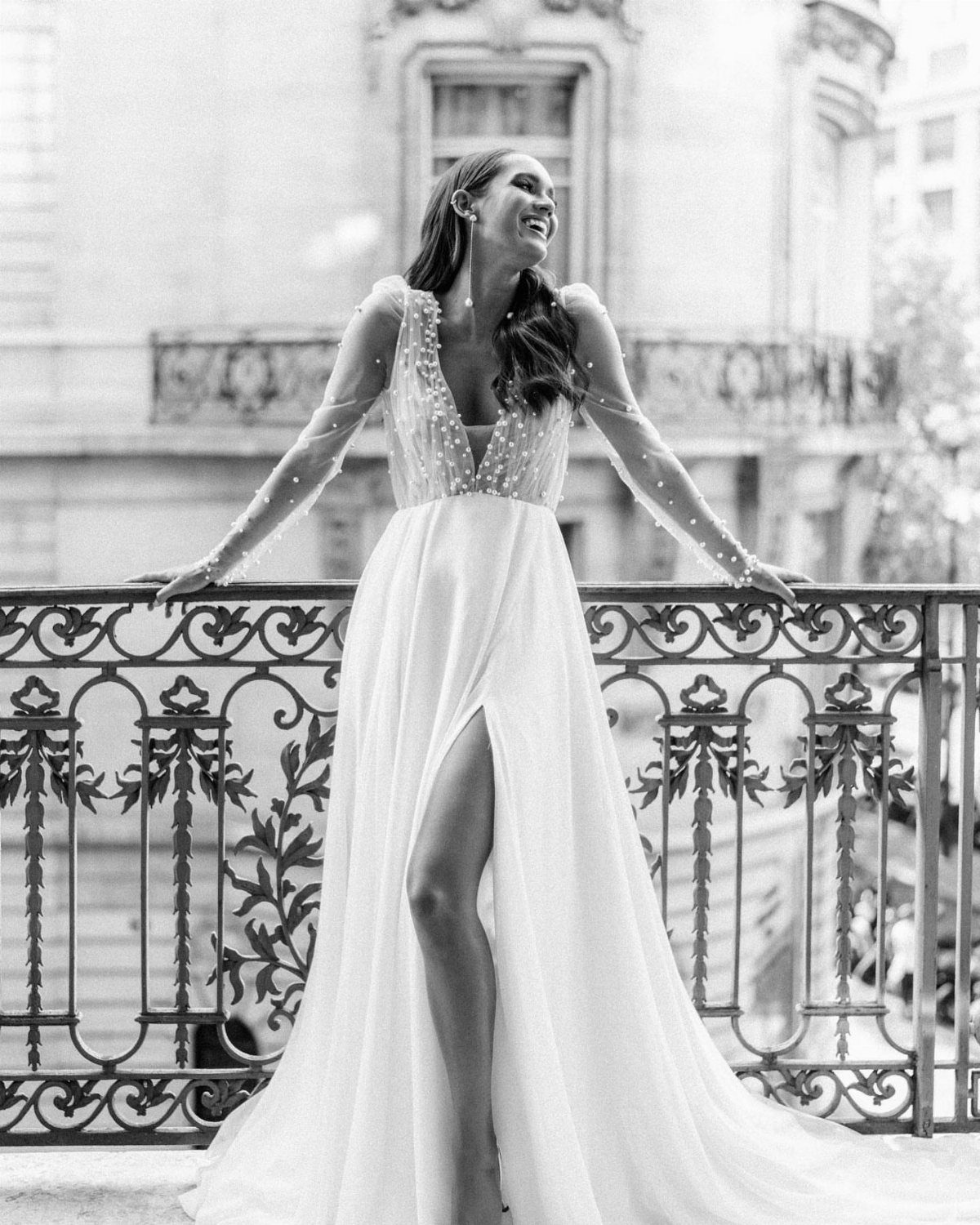 Art Deco Wedding Dress: 11 Styles From Beaded Trains to Choker Accents 2 A smiling woman in a beaded V-neck wedding dress stands on an ornate balcony.