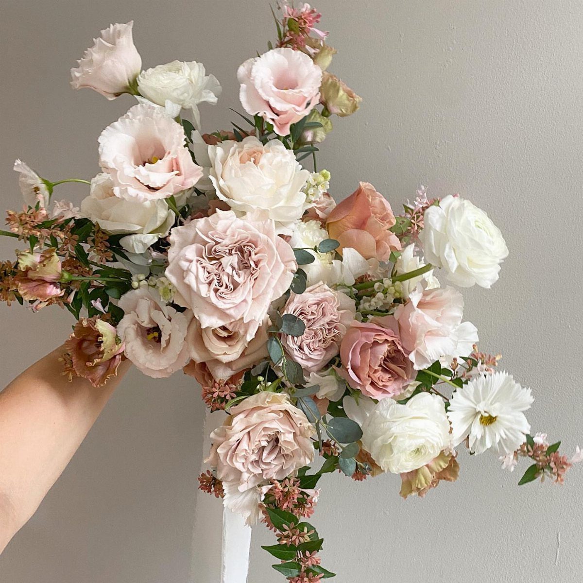A hand holding a large asymmetrical bouquet of pink and white roses with greenery.