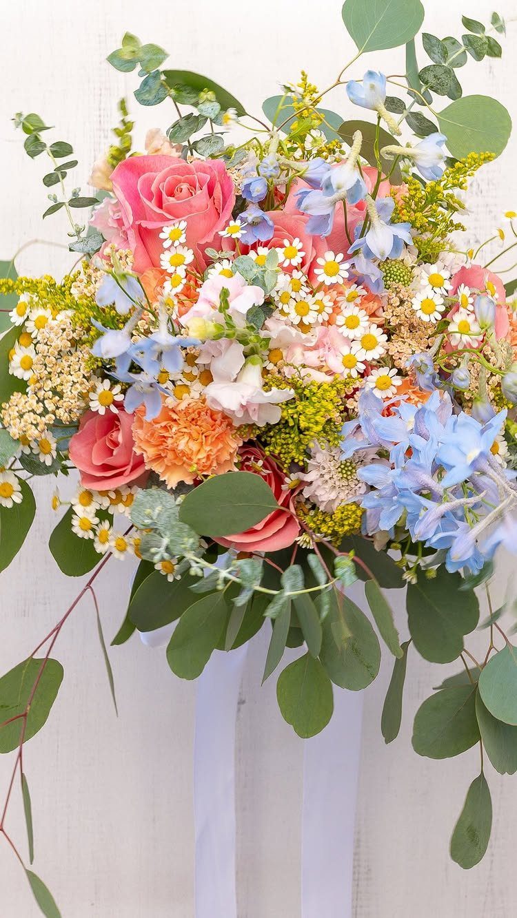 A colorful wedding bouquet with pink roses, blue delphiniums, and white daisies with eucalyptus leaves.