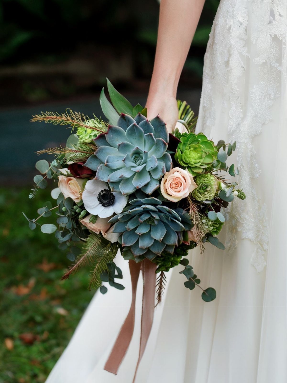 A bride holding a boho wedding bouquet with succulents, roses, and anemones with flowing ribbons.