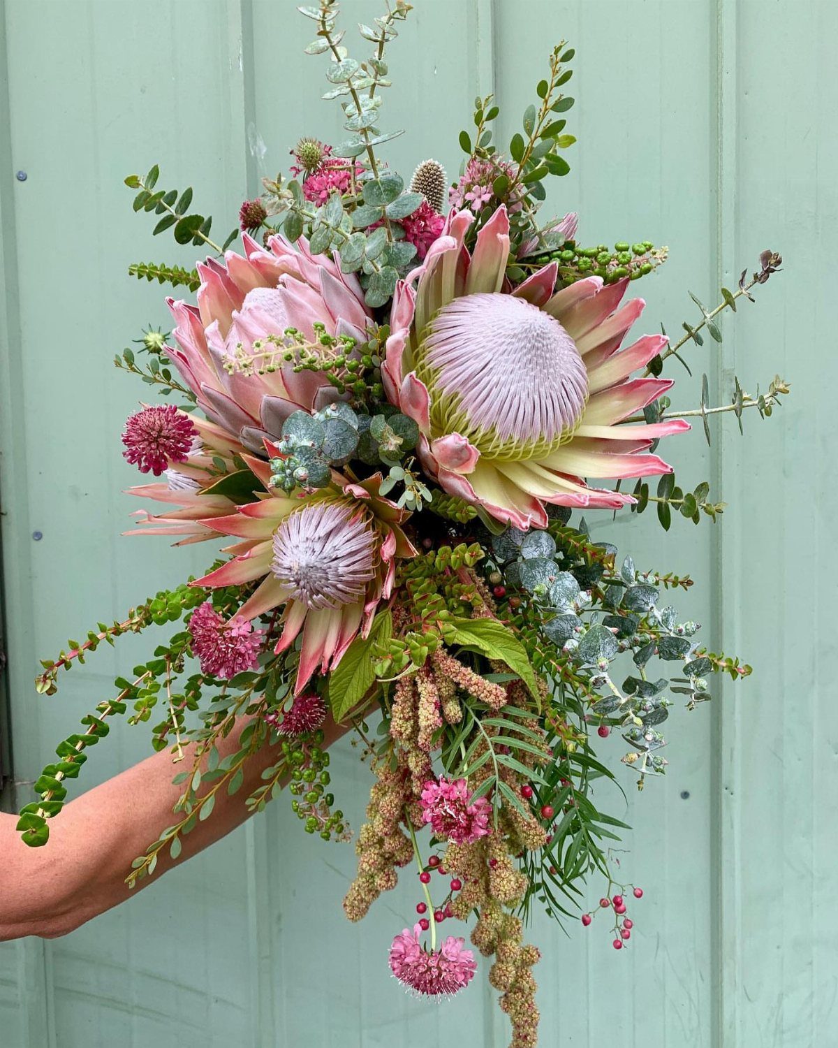 A lush wedding bouquet with pink proteas, eucalyptus leaves, and small pink wildflowers held against a green wall.