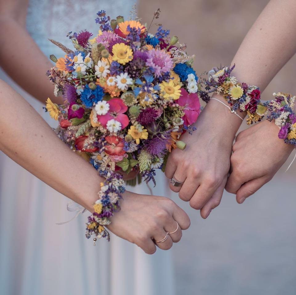 A vibrant bouquet of colorful wildflowers held by hands with matching floral wrist corsages.
