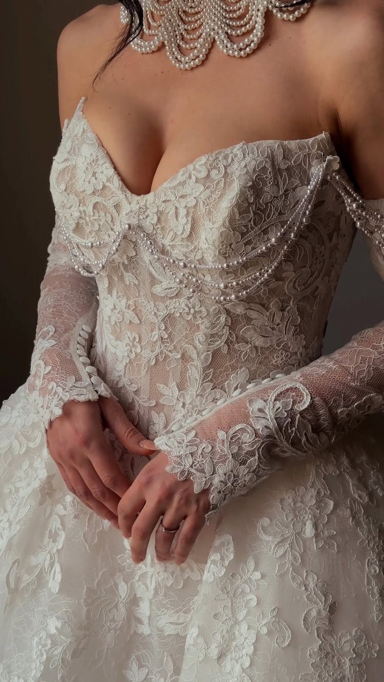 A person wearing an intricate white lace wedding dress with pearl embellishments and a pearl necklace.