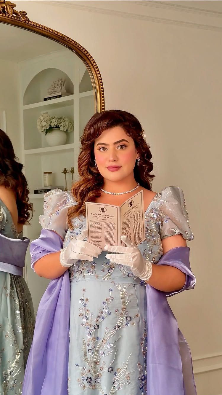 A woman in a light blue Regency-style gown with silver embroidery holding an open booklet.