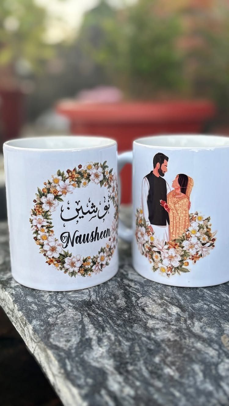 Two personalized white mugs with floral designs and a couple illustration on a stone ledge.