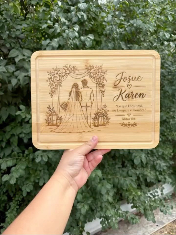 A hand holding a wooden cutting board engraved with a wedding couple illustration and names.