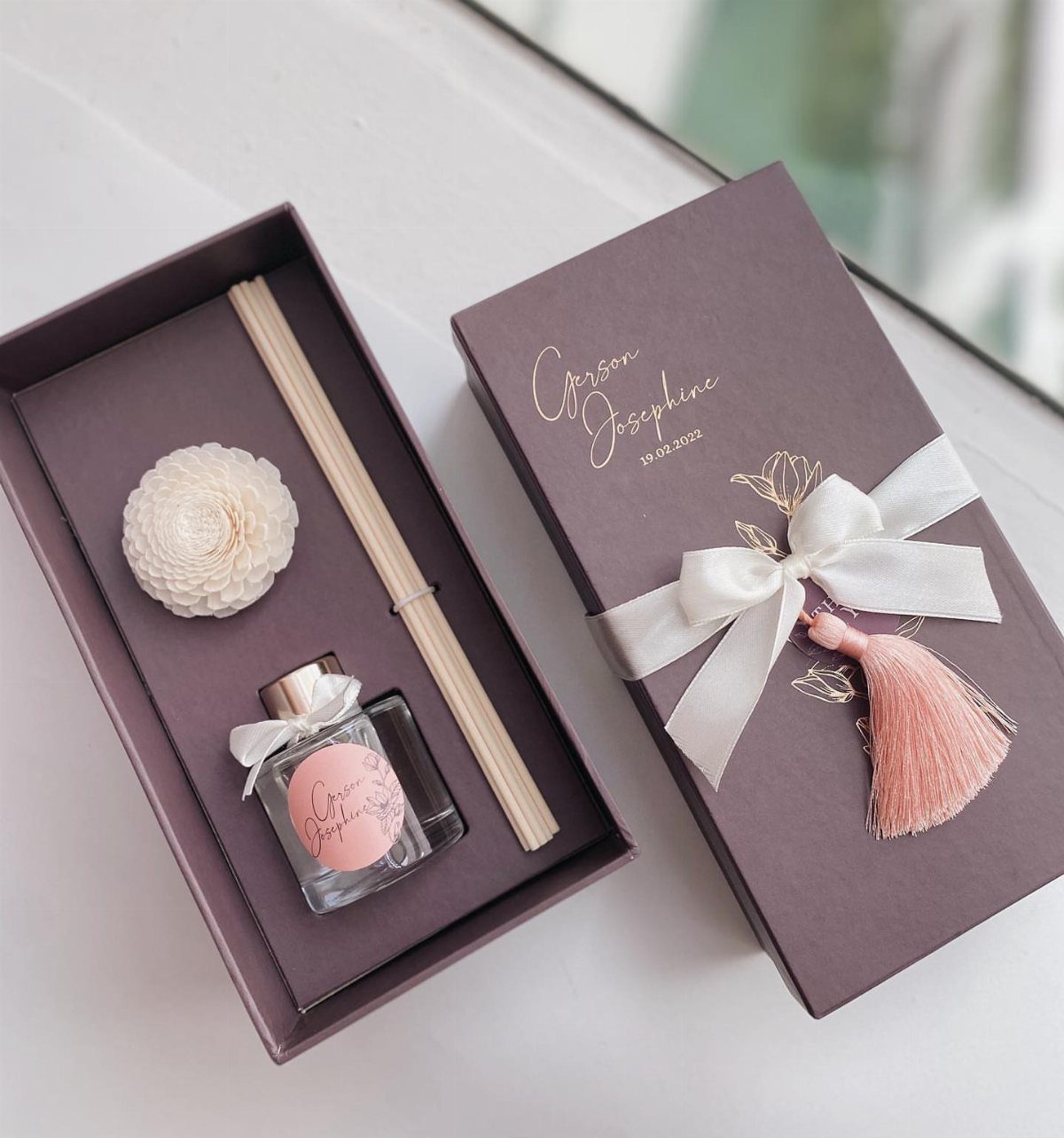 A reed diffuser gift set in a mauve box with a decorative white flower and pink tassel.