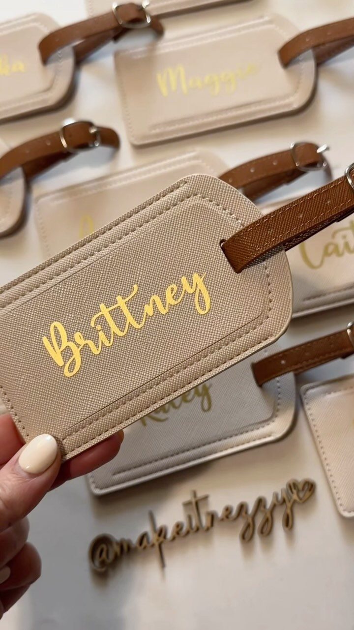 A hand holding a tan fabric luggage tag with the name Brittney printed in gold script.