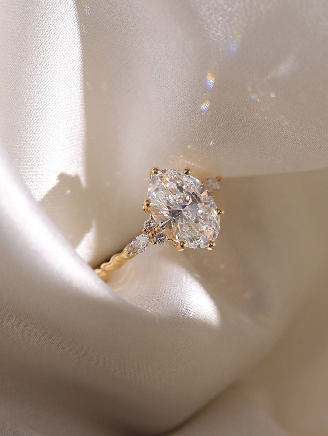 An oval cut diamond engagement ring with gold band resting on white silk fabric.