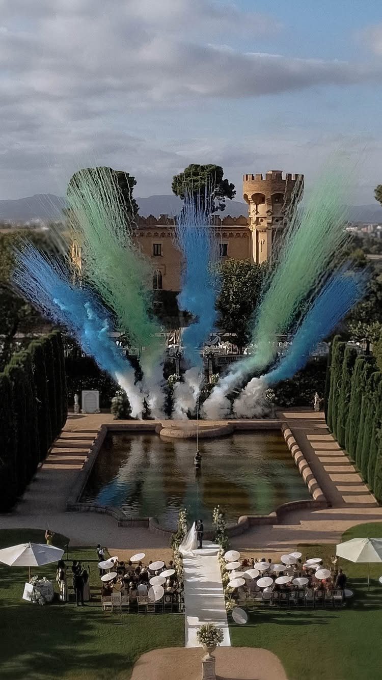 Wedding ceremony with colorful smoke plumes in front of a majestic castle and reflecting pool.