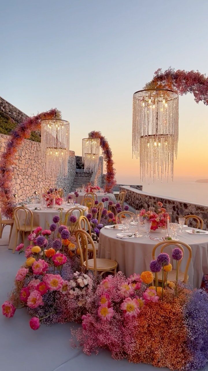 Outdoor wedding reception tables decorated with colorful flowers and crystal chandeliers under a sunset sky.