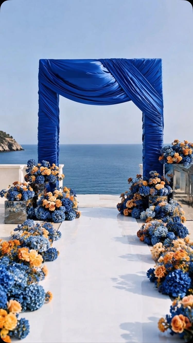 Blue fabric wedding arch and floral arrangements lining a white aisle overlooking the ocean.