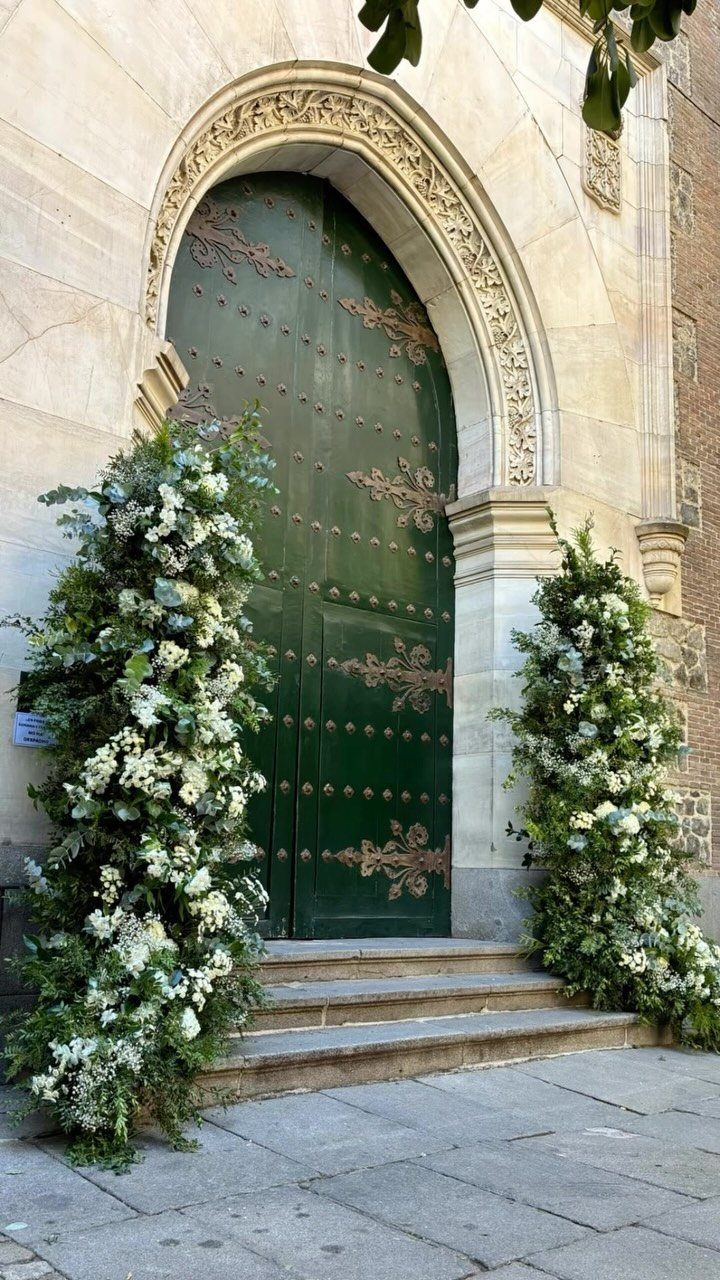 Large white floral arrangements flanking a grand arched green wooden door in a stone building.