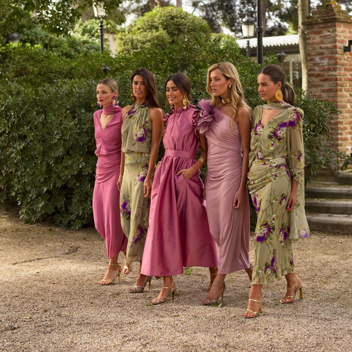 Five women posing in elegant formal dresses and high heels in a lush garden setting.