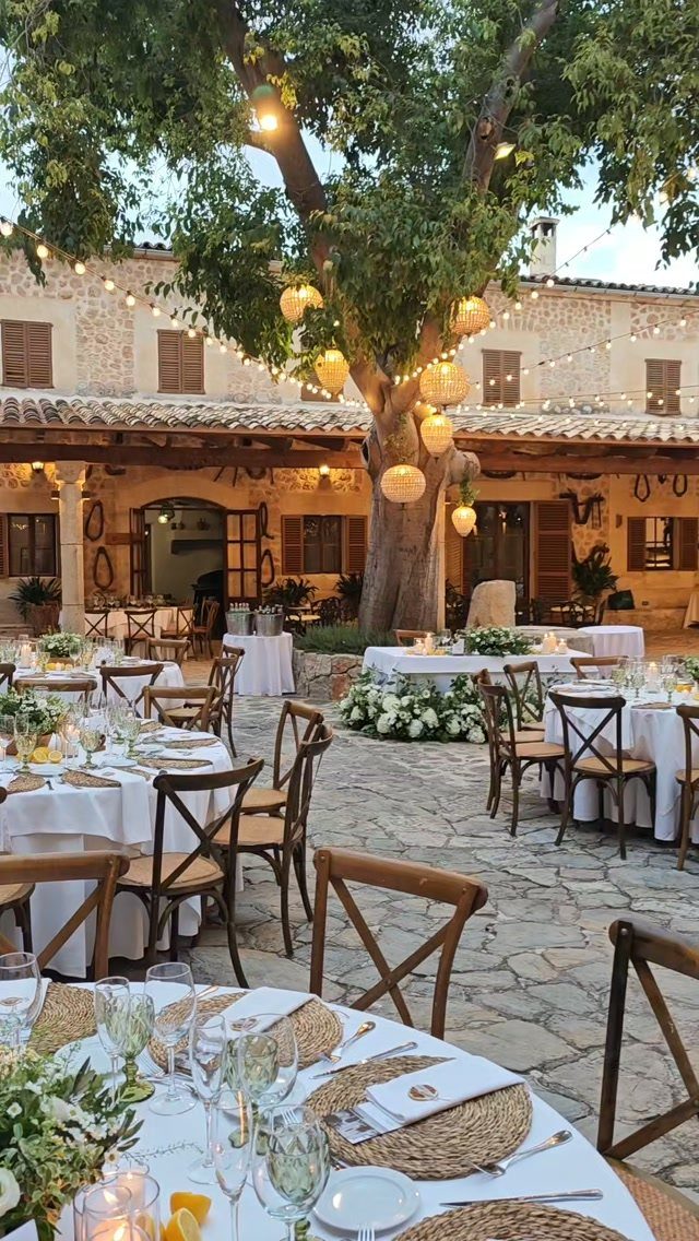 Dining tables set in a stone courtyard under a large tree with hanging lanterns.