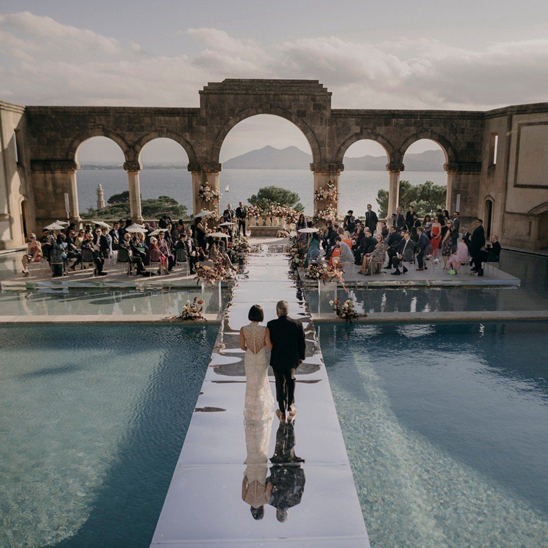 A bride and groom walk across a white bridge over a pool toward a wedding ceremony.