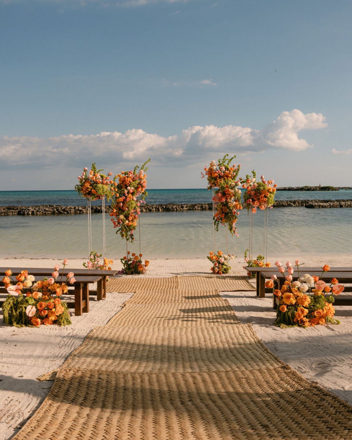 A beach wedding ceremony setup with a jute aisle runner and orange floral arrangements by the sea.