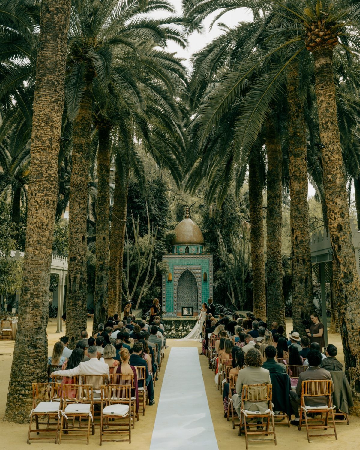 Wedding guests seated in rows facing a turquoise altar under tall palm trees in an outdoor setting.