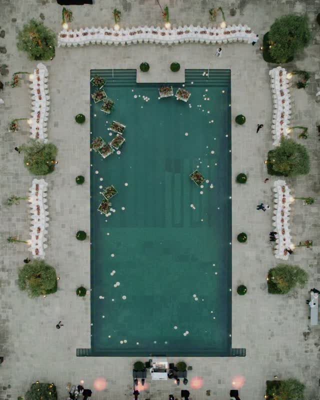 Aerial view of a wedding reception with white tables arranged around a turquoise swimming pool.