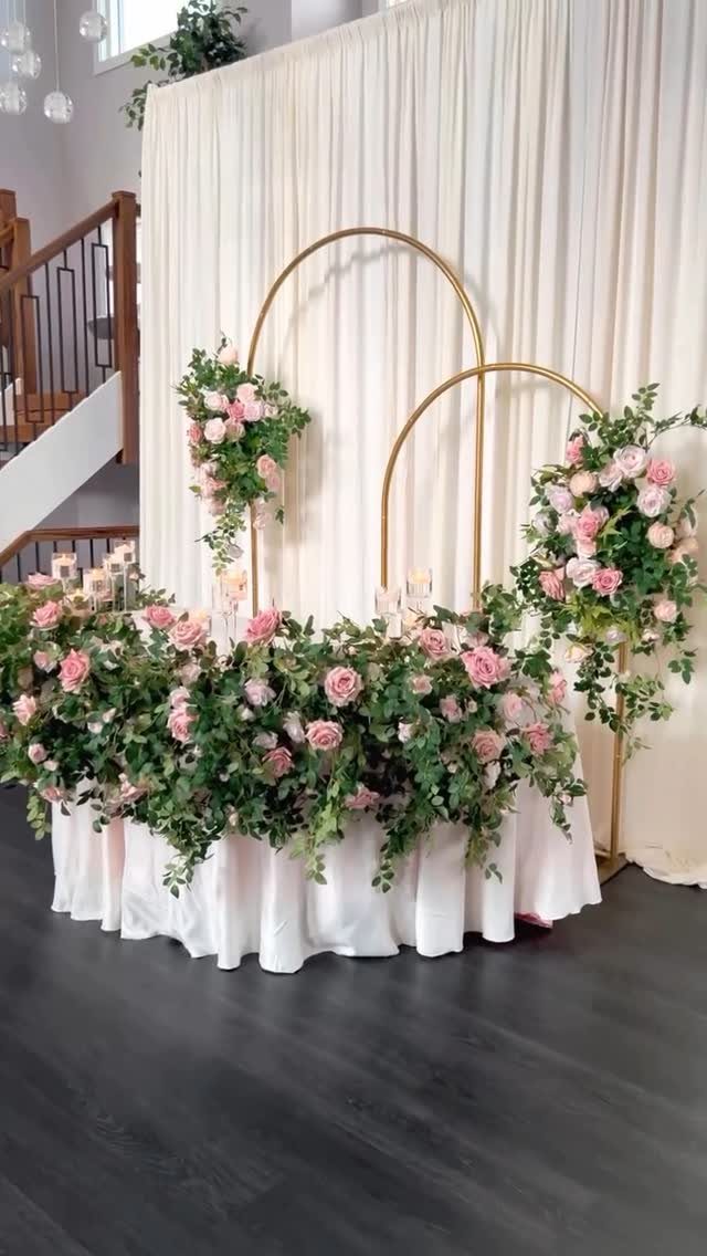 Flowers for Sweetheart Table: 6 Ideas for a Focal Point 2 Pink rose floral arrangements decorating a sweetheart table with gold arch backdrops and white drapery.