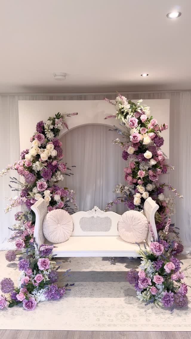 Flowers for Sweetheart Table: 6 Ideas for a Focal Point 6 A white loveseat framed by lush arrangements of purple, pink, and white flowers against a white backdrop.