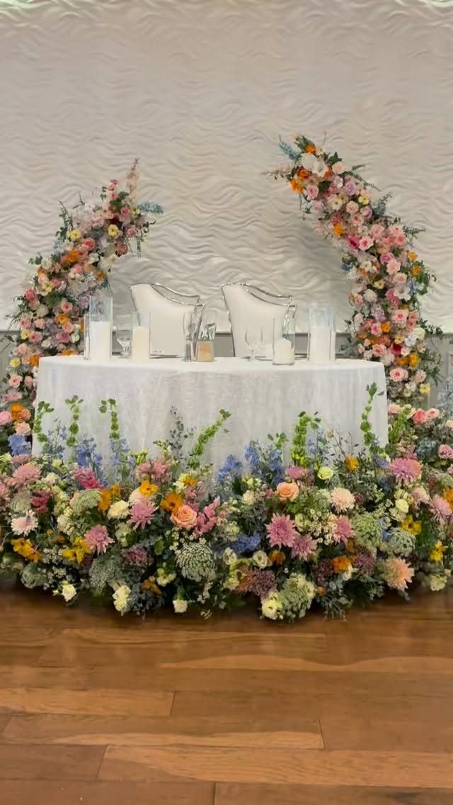 Flowers for Sweetheart Table: 6 Ideas for a Focal Point 5 A wedding sweetheart table decorated with vibrant floral arches and a thick flower arrangement along the base.