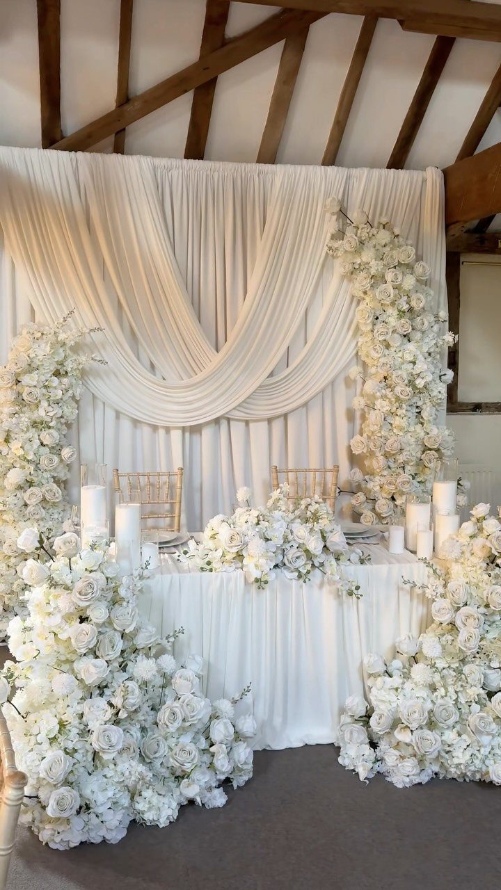 Flowers for Sweetheart Table: 6 Ideas for a Focal Point 4 White floral arrangements surround a decorated sweetheart table with draped fabric backdrop and candles.