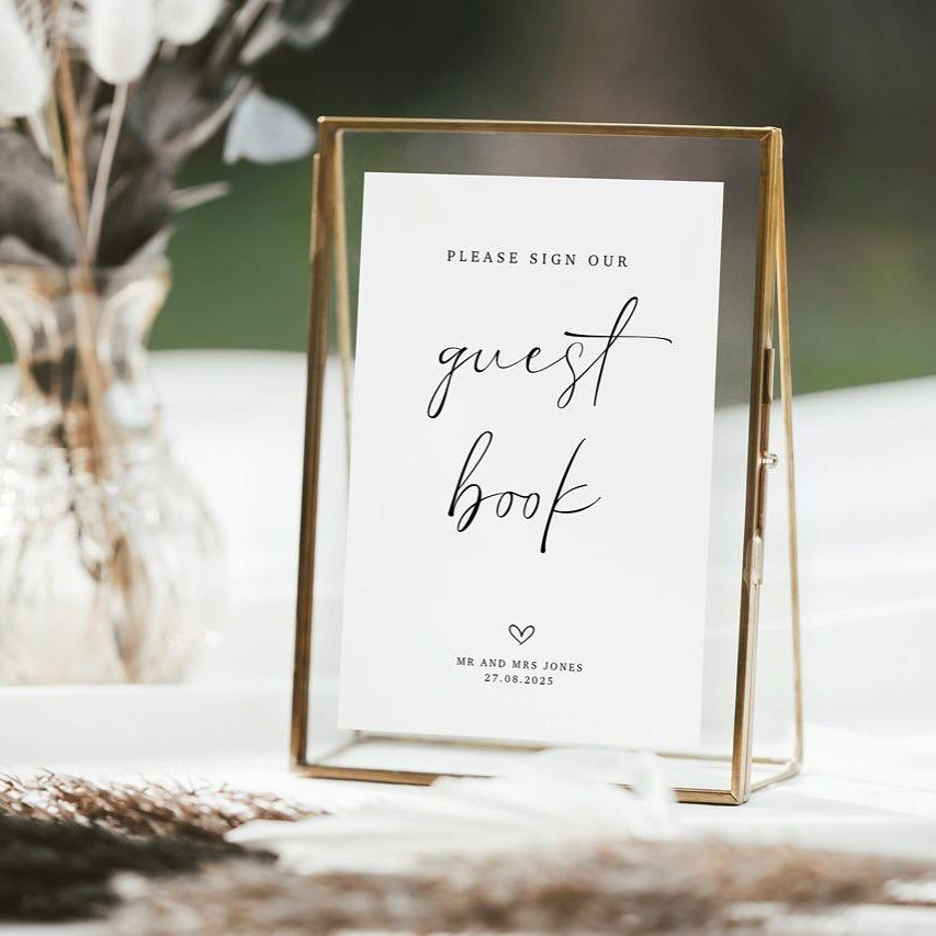 A white guest book sign in a gold glass frame sits on a wedding table.