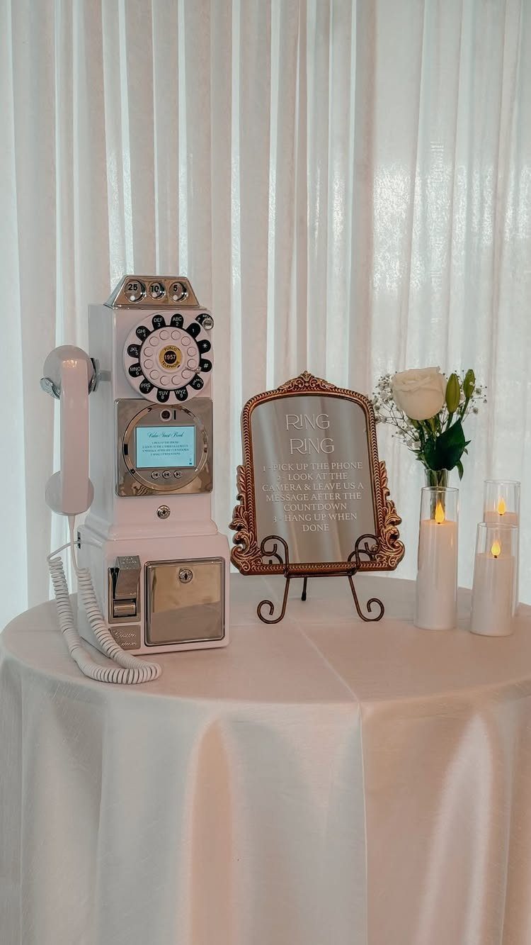 A white rotary phone audio guest book on a white table with a gold frame sign and flowers.