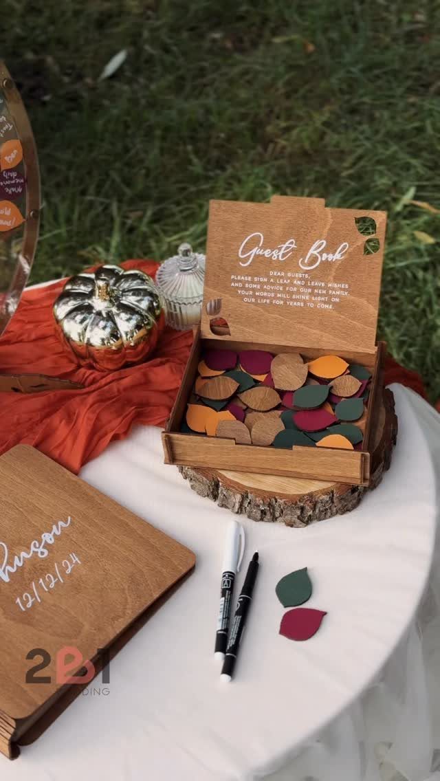 Wooden guest book box filled with colorful leaf shapes and a large wooden book on a table.