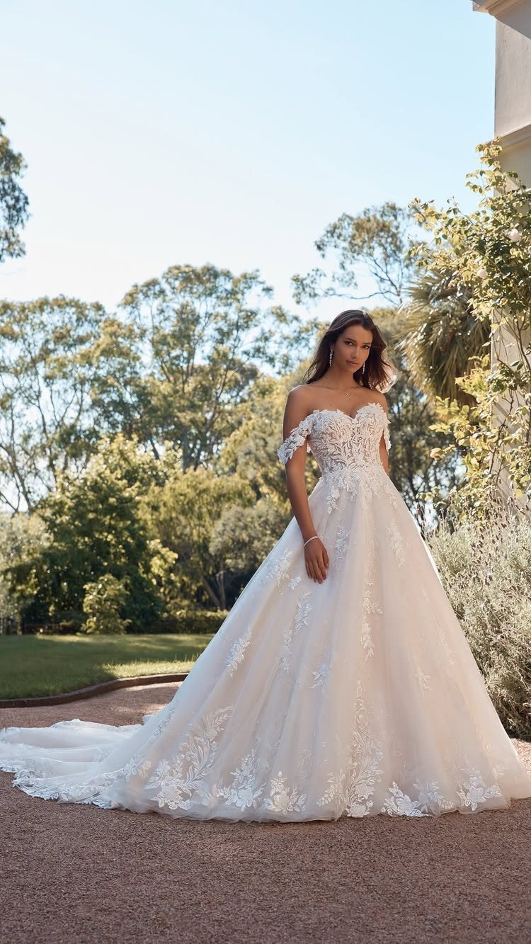 A woman wearing a white lace off-the-shoulder ballgown wedding dress standing in a sunny garden.