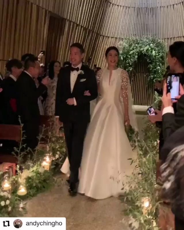 A bride in a white long-sleeved gown and a groom walk down a floral wedding aisle.