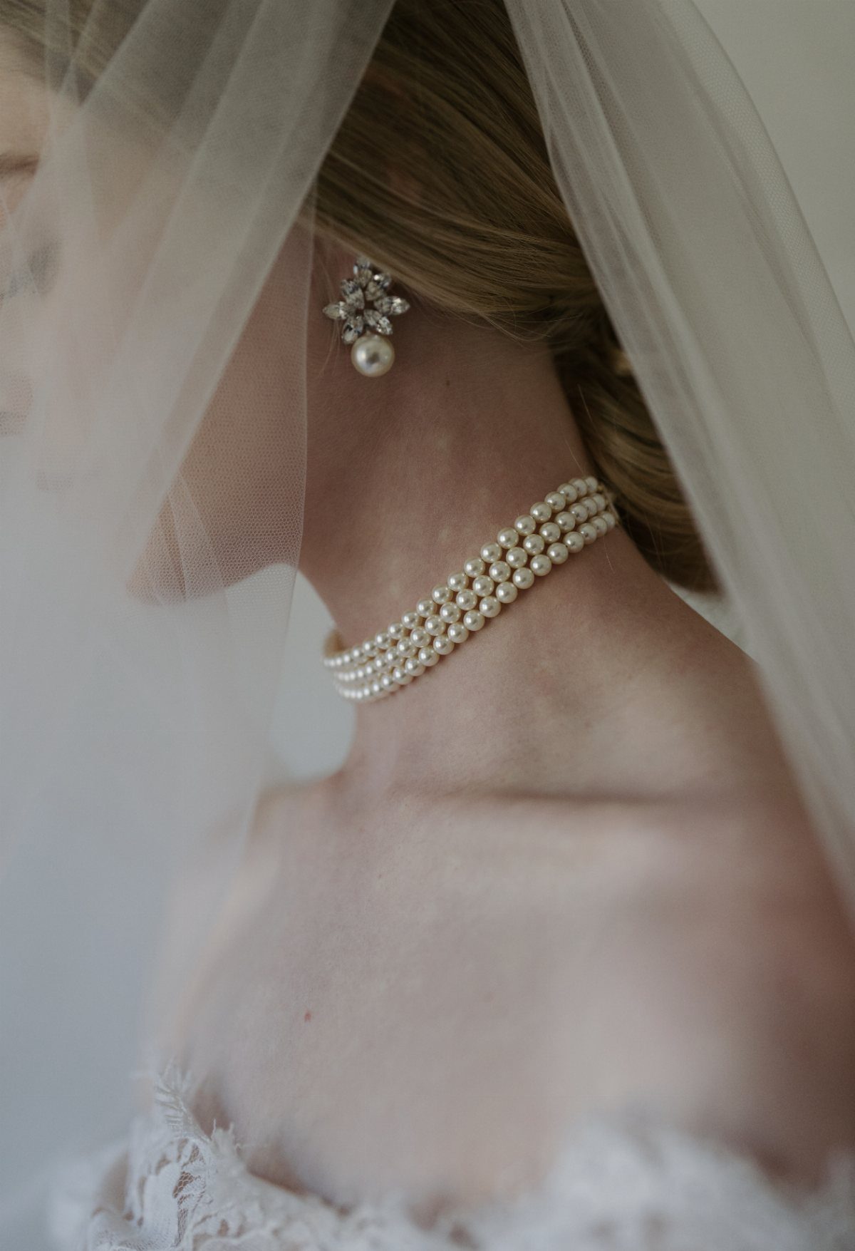 A woman wearing a sheer white tulle veil, pearl choker, and pearl earring.