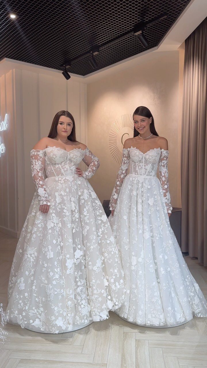 Two women posing in elegant, off-the-shoulder white lace wedding dresses indoors.