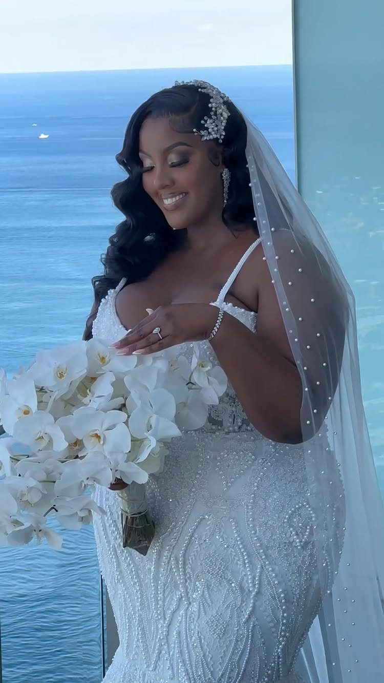A smiling bride in a beaded white wedding dress holding white orchids by the ocean.