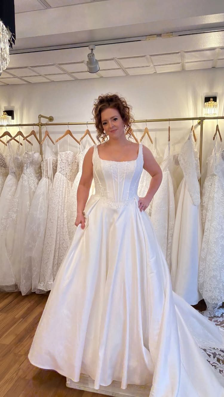A woman wearing a white satin wedding dress with a square neckline in a bridal shop.