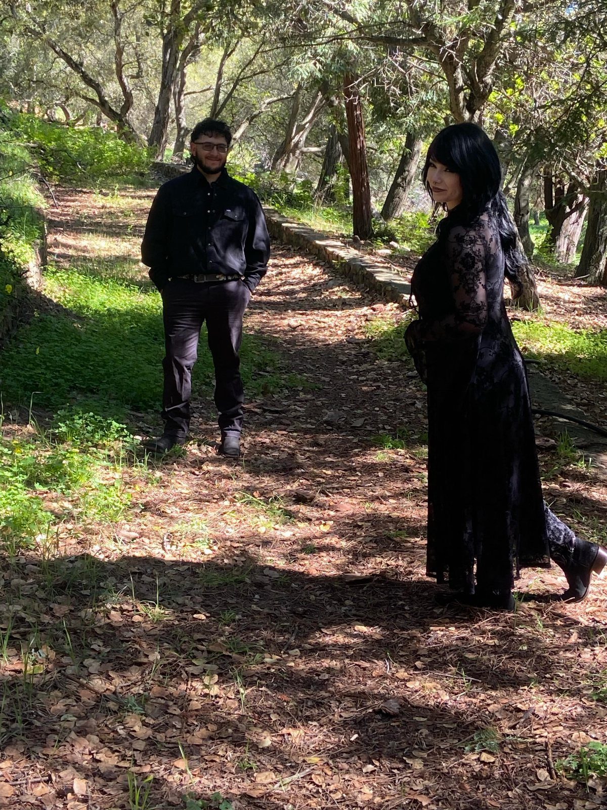 A woman wearing a long black lace dress standing on a sunlit forest path.