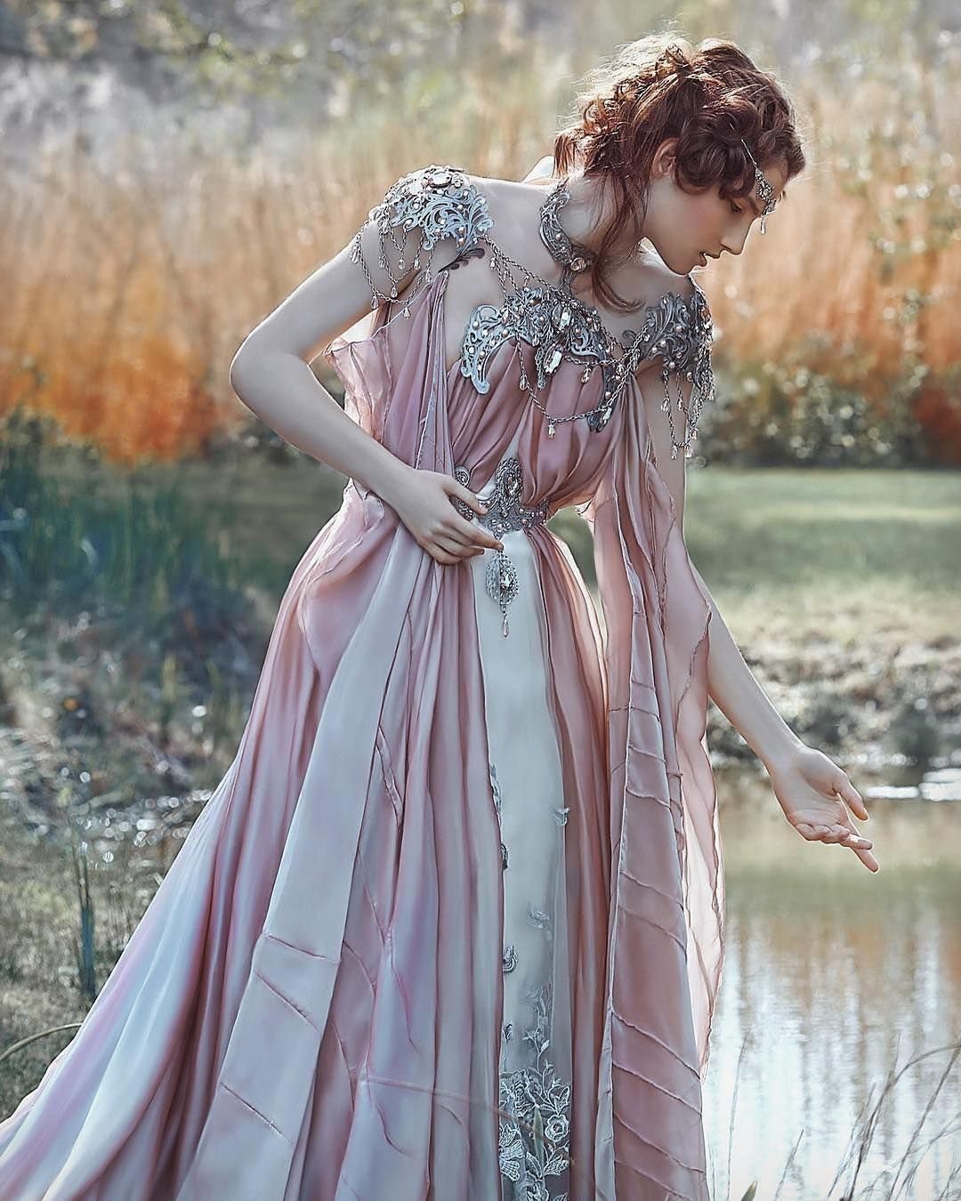 A woman wearing a dusty rose and white gown with silver embroidery standing outdoors by water.
