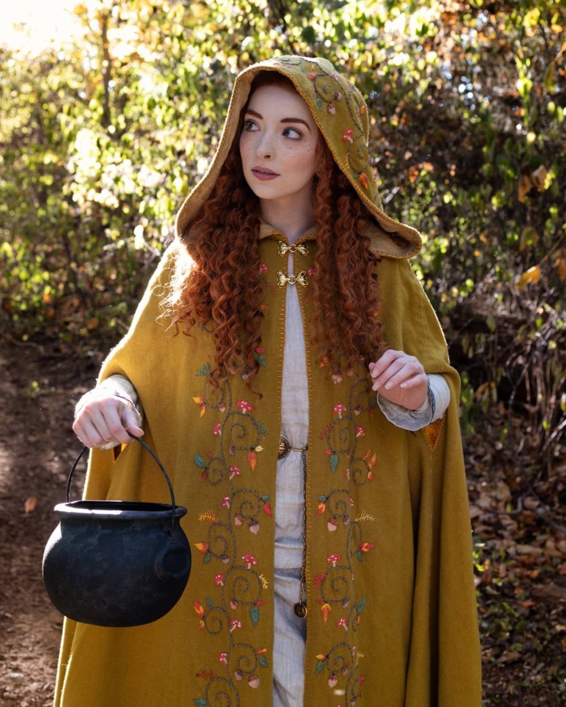 Woman with long red hair wearing an embroidered mustard yellow hooded cloak in a forest.