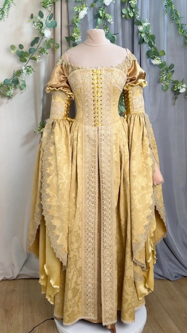 A gold damask lace gown with a corset bodice and flared sleeves on a mannequin.