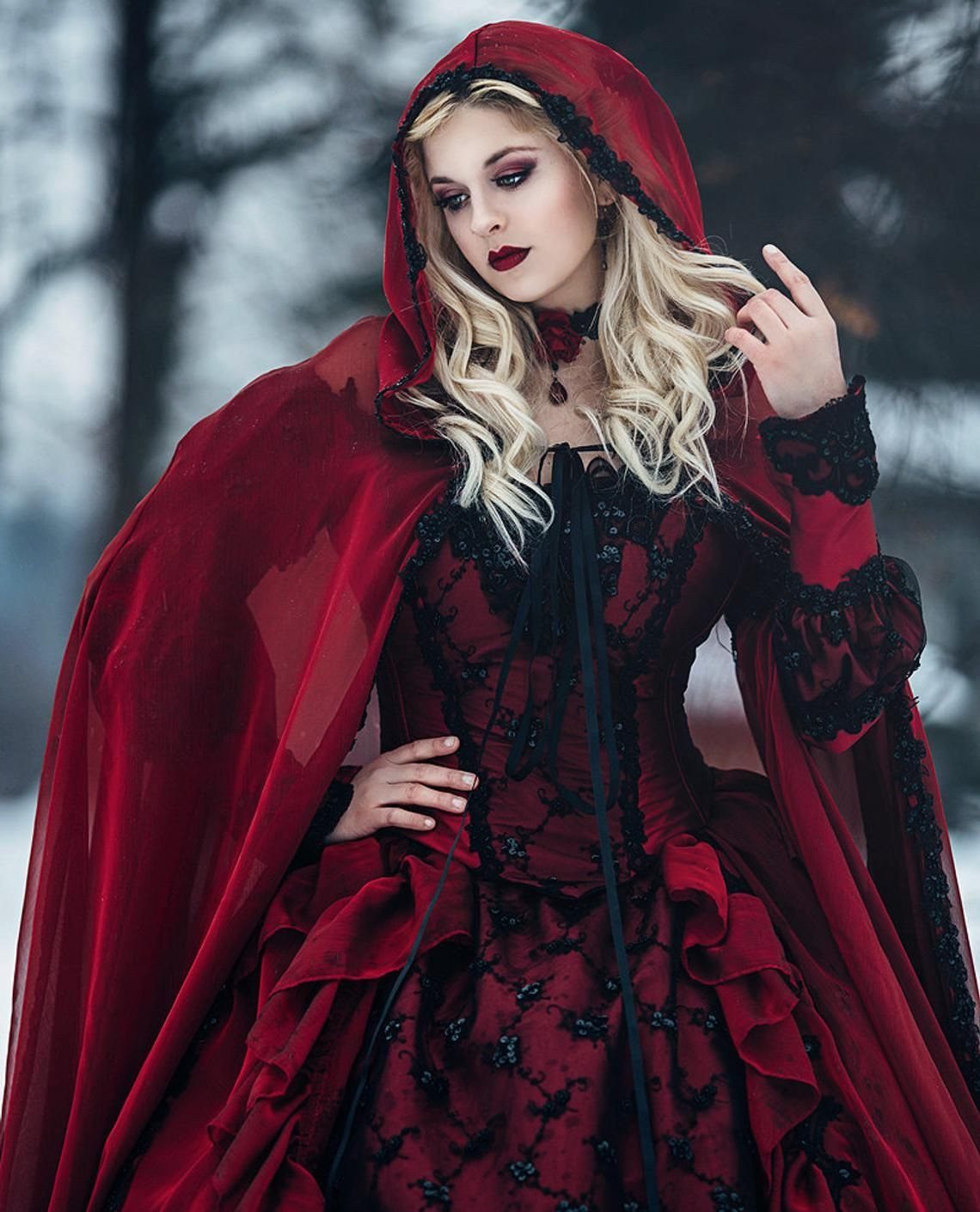 A blonde woman wearing a red lace gown with a matching hooded cloak in the snow.