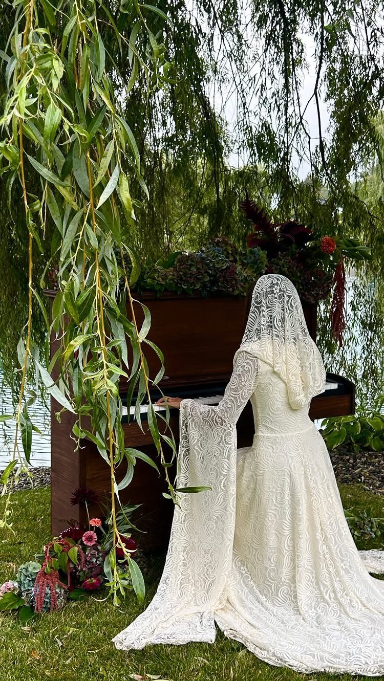 A person in a white lace gown with a long hooded sheer cape standing by a piano.