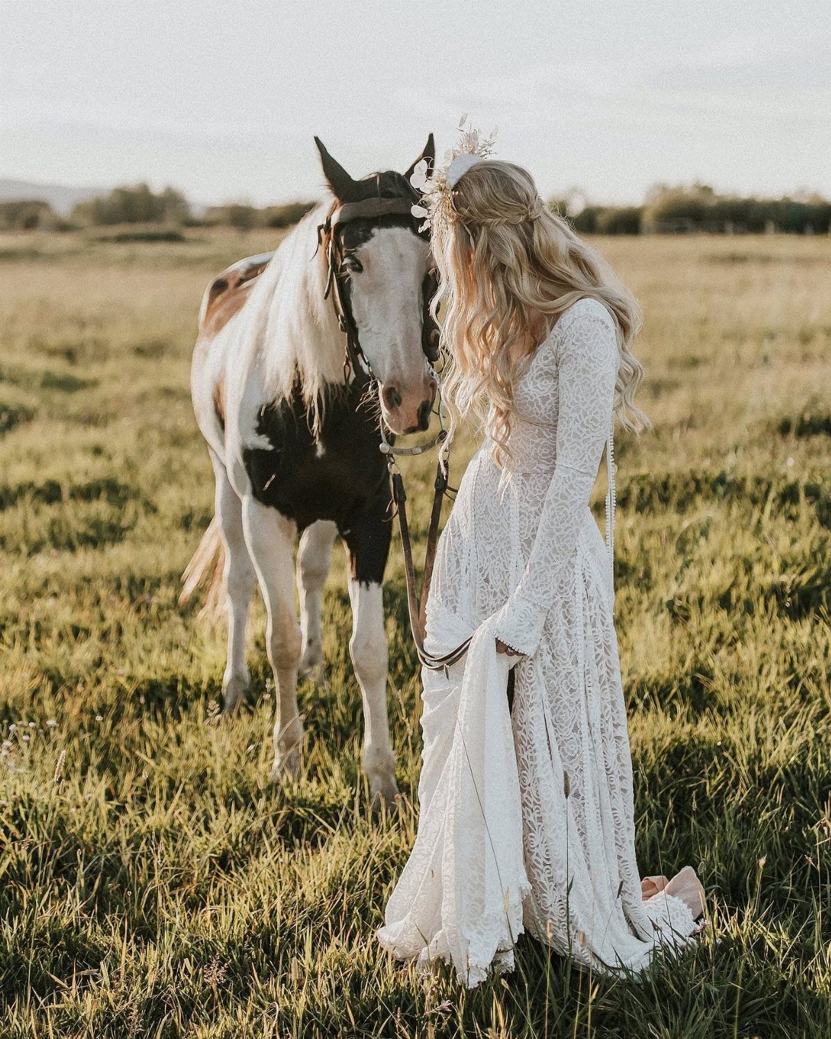 A woman in a white lace maxi dress and floral crown stands beside a horse in a field.